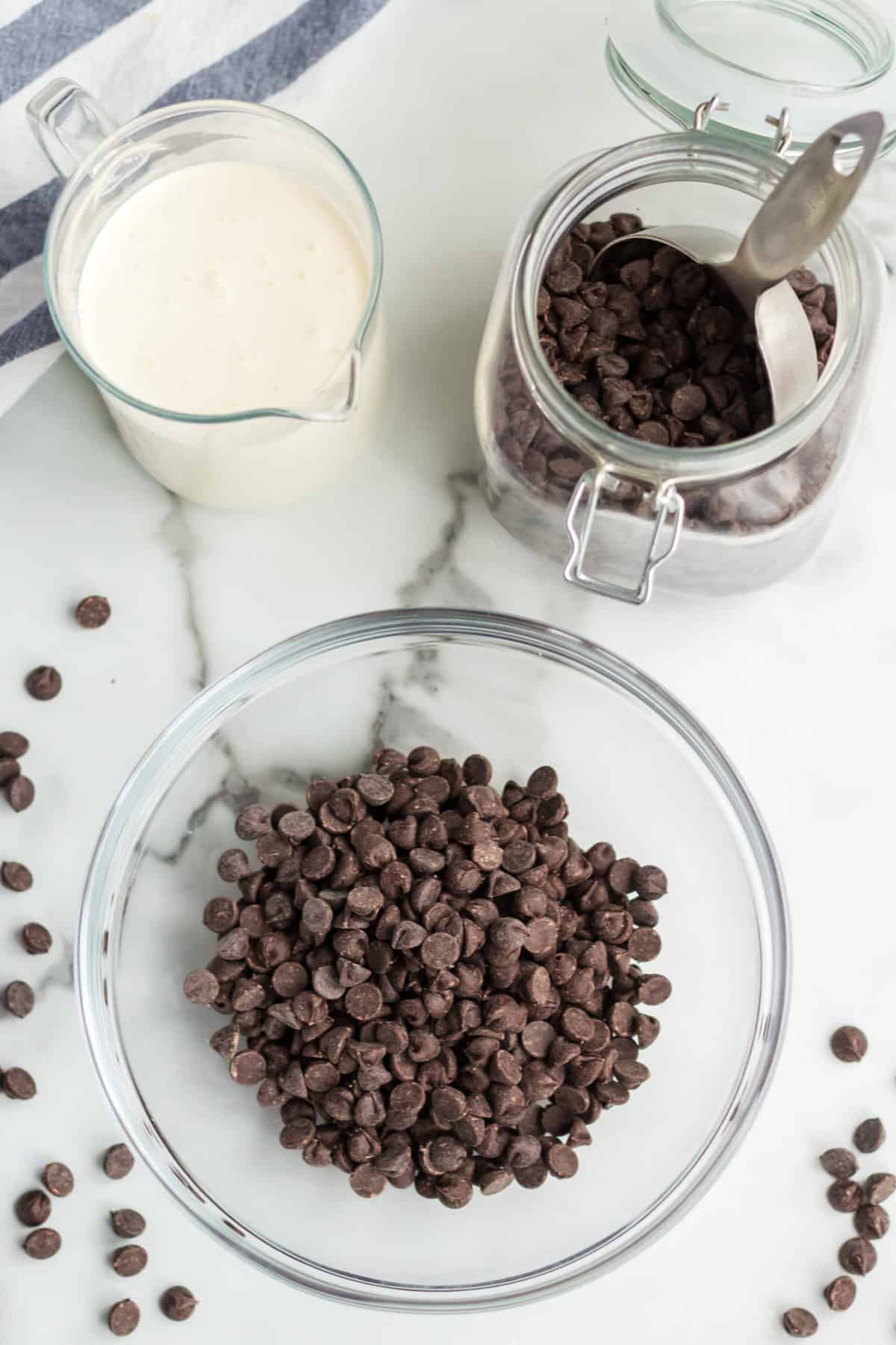 Picture of all the ingredients needed for the recipe including a glass bowl with chocolate chips, a small jug of heavy whipping cream, and a jar with extra chocolate chips with a measuring cup in it. 