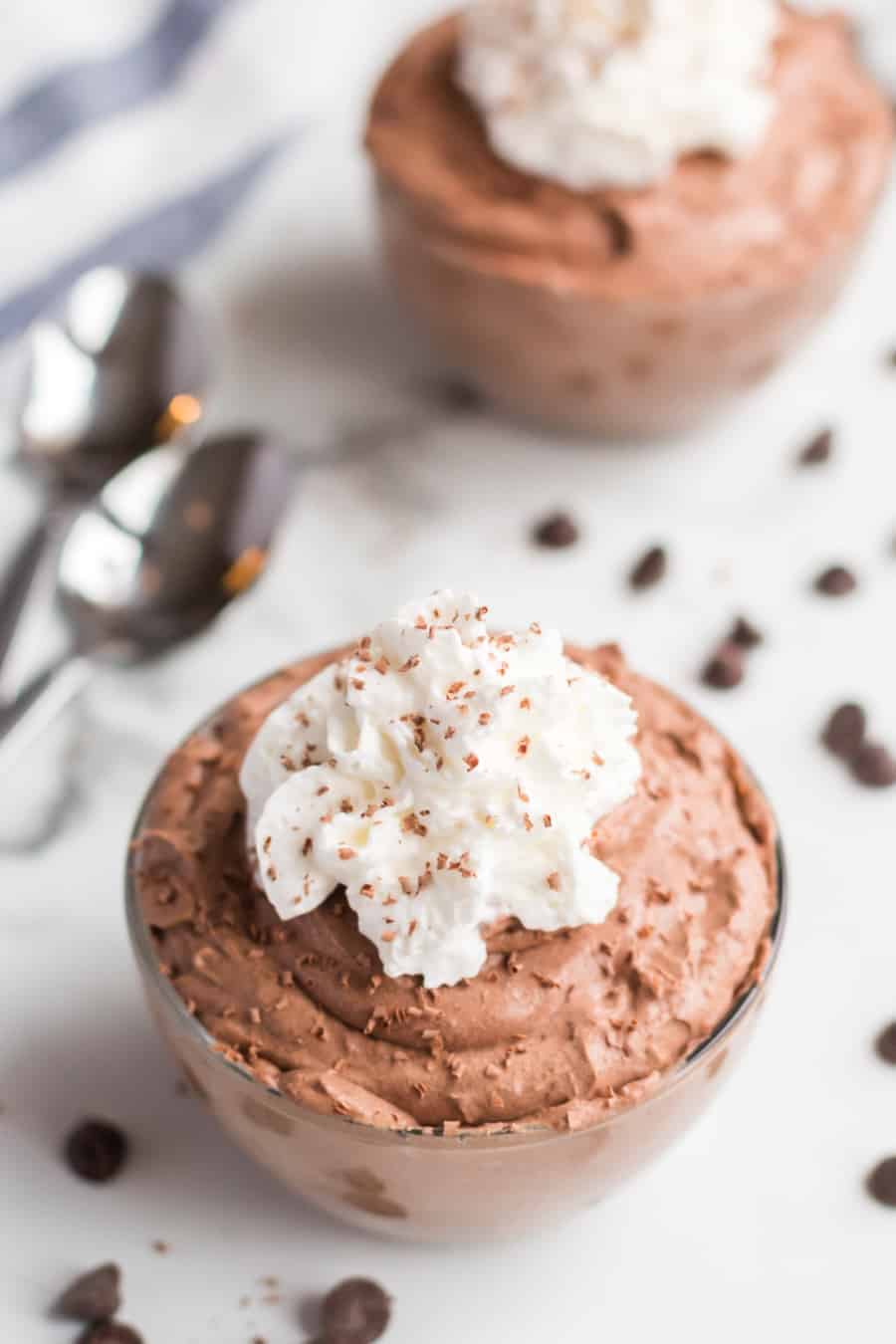 Small glass serving bowl filled with chocolate mousse that's topped with whipped cream and chocolate shavings, there are two spoons and another small bowl of mousse in the background. 