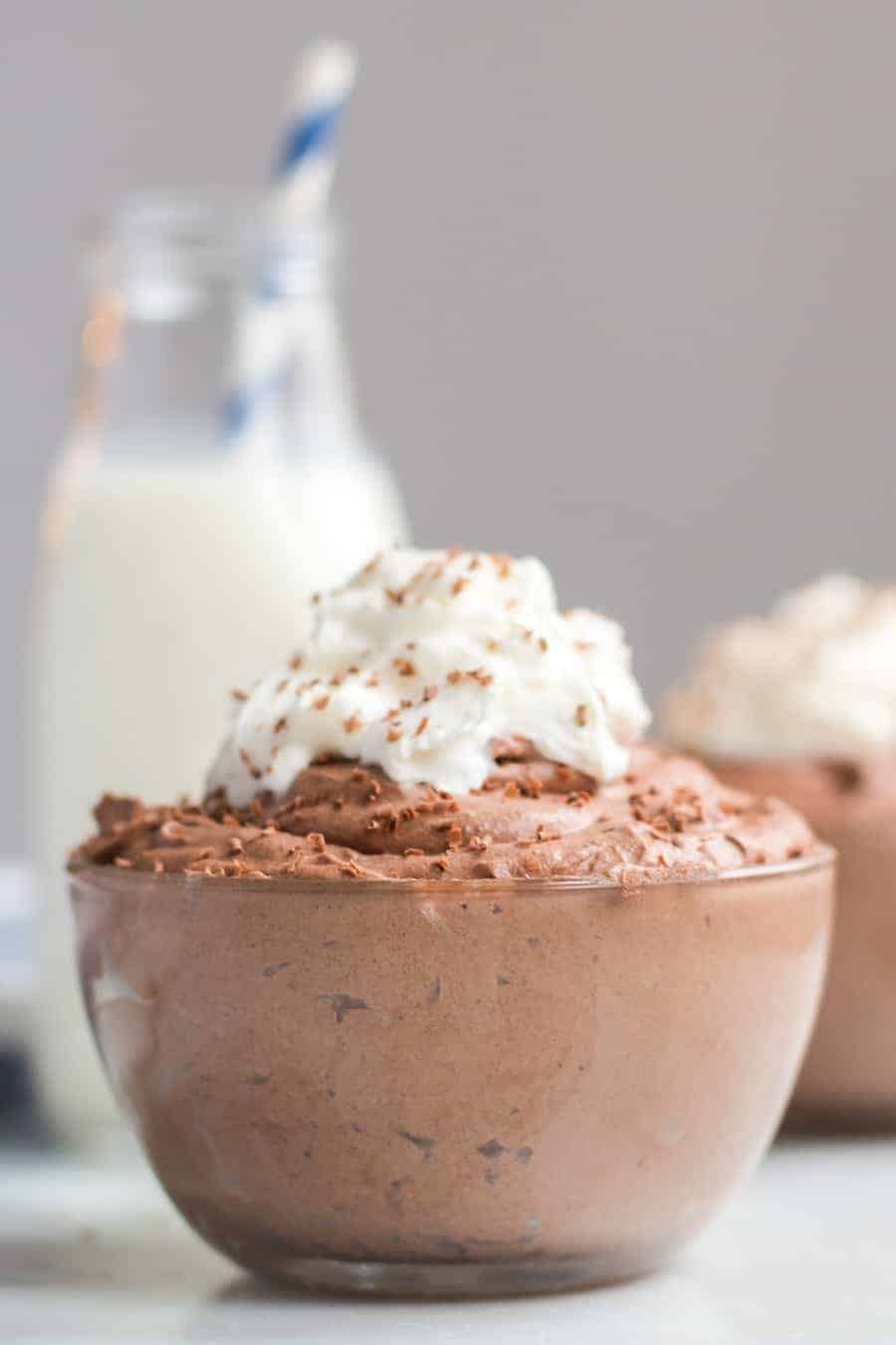Small glass bowl filled with chocolate mousse and topped with whipped cream and chocolate shavings with a bottle of milk and another bowl of mousse in the background. 