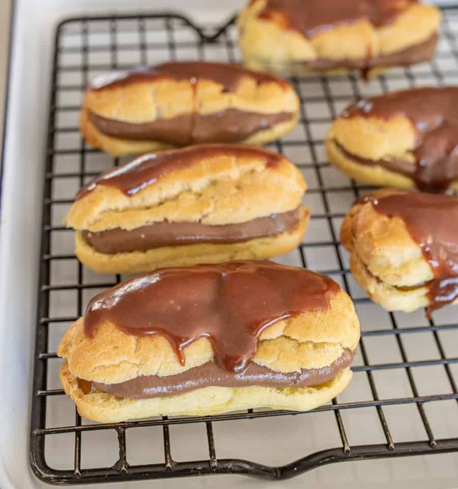 Finished eclairs stuffed with chocolate custard and topped with chocolate icing sitting on a black cooling rack. 