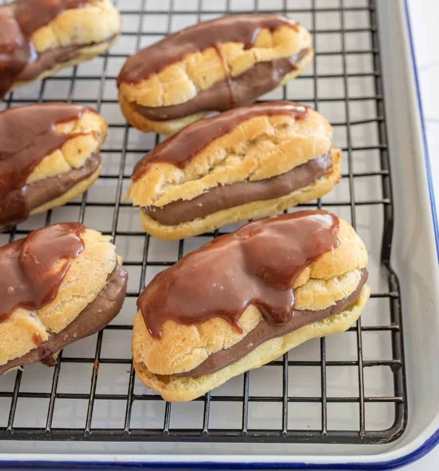 Finished eclairs stuffed with chocolate custard and topped with chocolate icing sitting on a black cooling rack. 