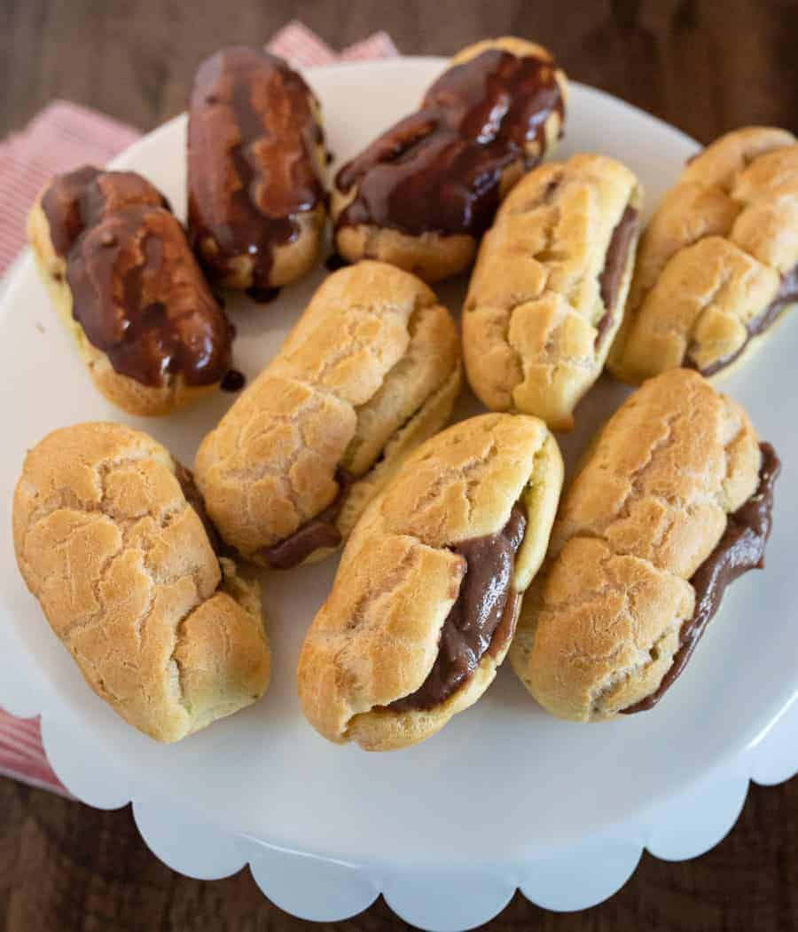 Nine finished eclairs on a white plate stuffed with chocolate custard, some have chocolate icing and some are plain. 