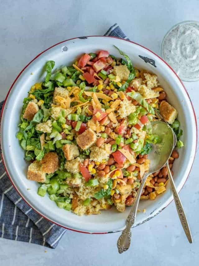 A large bowl filled with layered cornbread salad ingredients including chopped lettuce, tomatoes, shredded cheese, croutons, pinto beans, corn, and green onions, with two serving spoons beside the bowl.