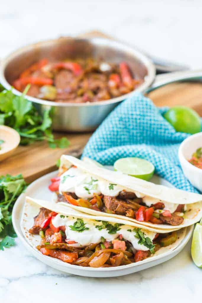 flour tortillas with steak and peppers with lime on a white plate with fajita mix in the background
