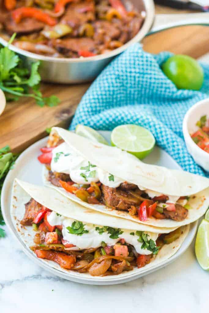 flour tortillas with steak and peppers with lime on a white plate with fajita mix in the background