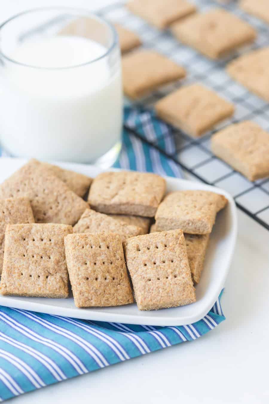 simple homemade graham crackers on white dish with a blue towel and cooling rack with a glass of milk
