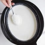 white slow cooker yogurt in a black slow cooker or crock pot with hand holding spoon dipped in yogurt.