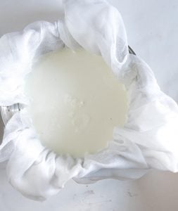 A bowl lined with white cheesecloth holds thick, white slow cooker yogurt being strained on a light surface.