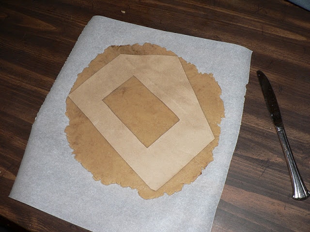 Gingerbread dough rolled out on parchment paper with a house-shaped parchment stencil on top