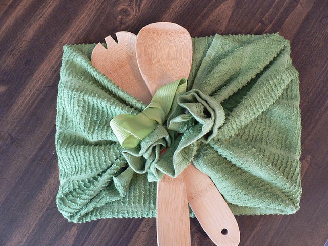 A square gift wrapped in a green tea towel with two wooden spoons