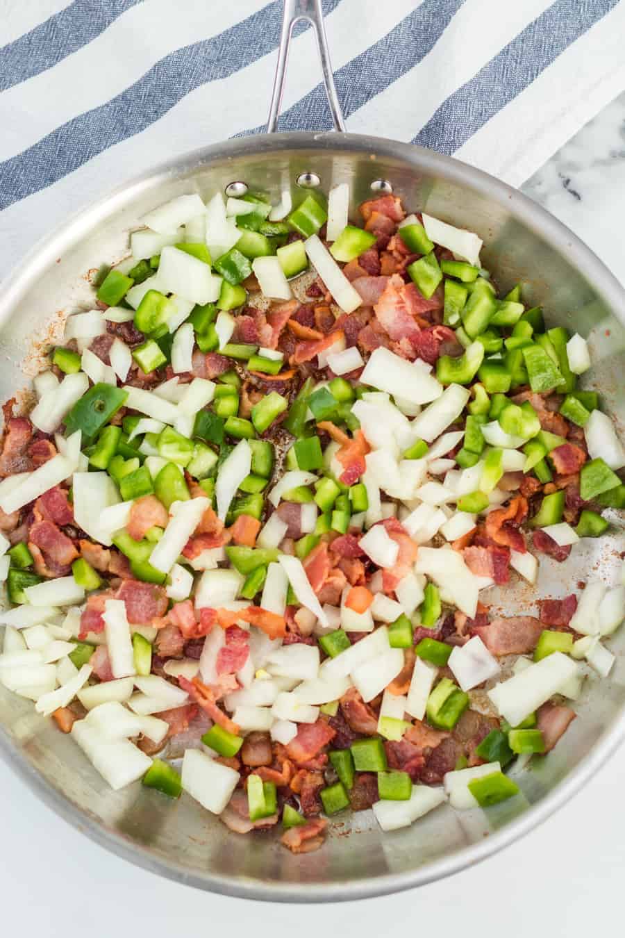 Stainless steel skillet with cooked bacon and chopped onion and green bell pepper ready to cook into a pasta sauce.