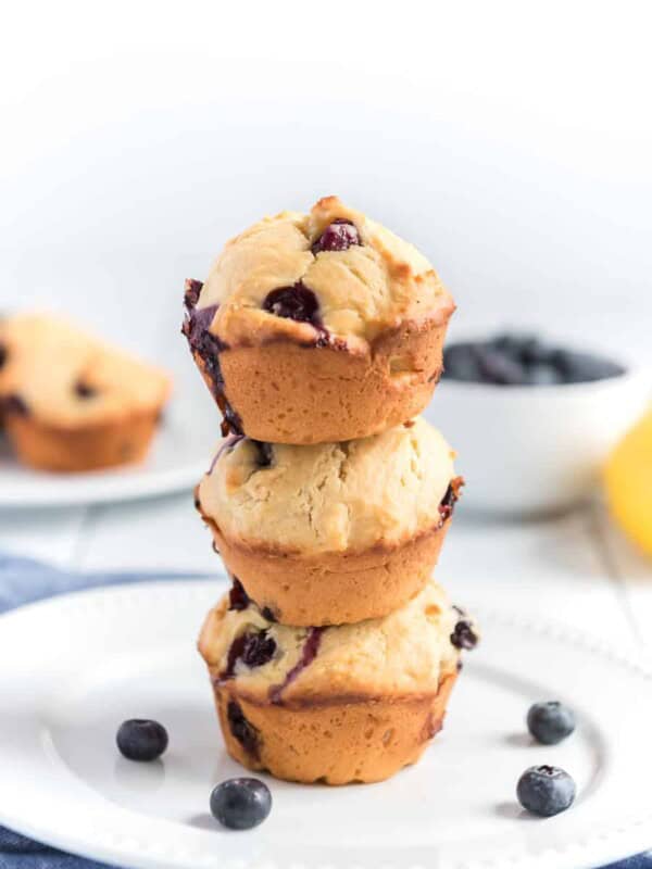 A stack of three golden-brown lemon blueberry muffins sits on a white plate, with a few blueberries scattered around. A bowl of blueberries and a partially visible muffin are in the blurred background.