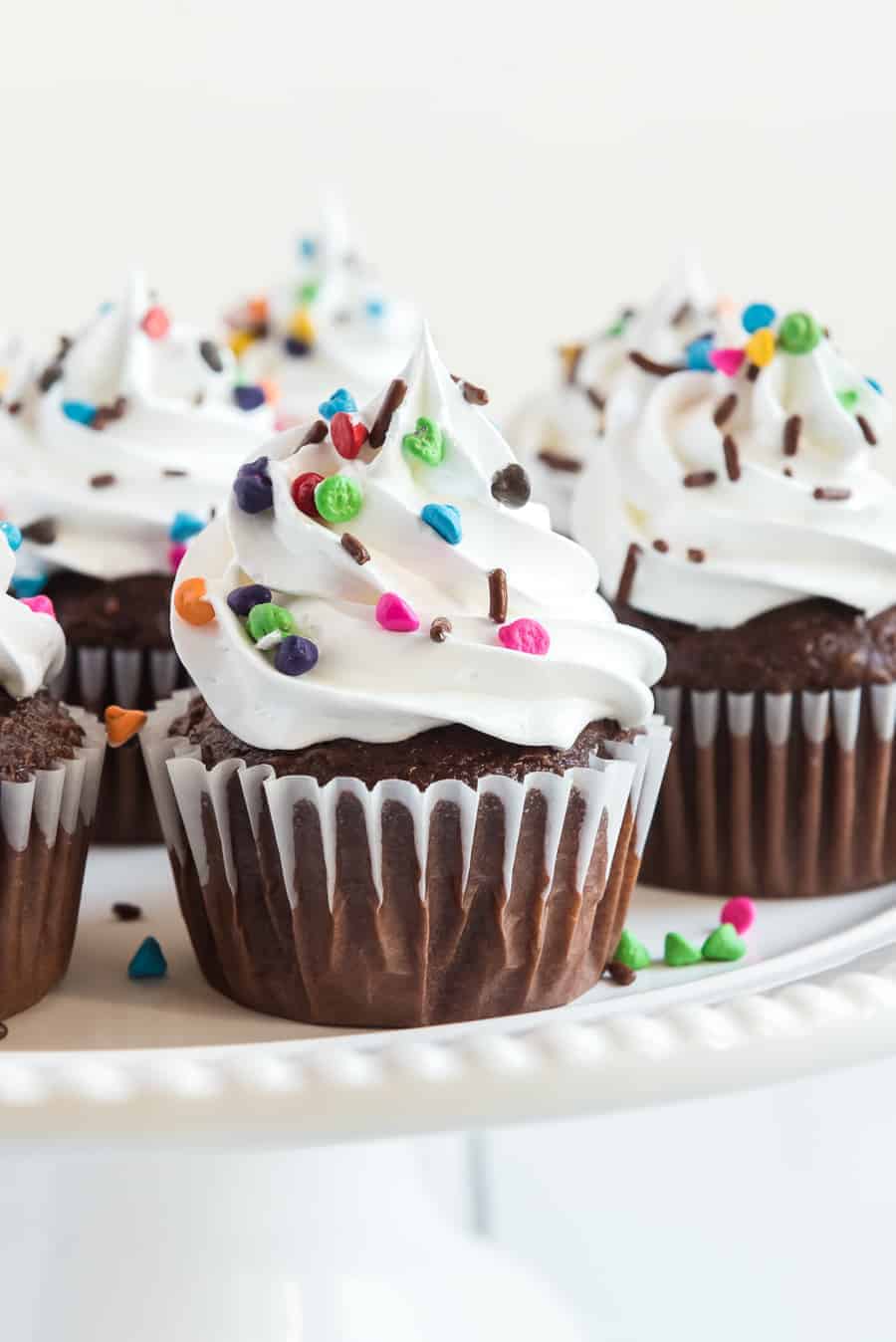 close up image of chocolate cupcakes with white frosting and multi-colored sprinkles all on a white plate.