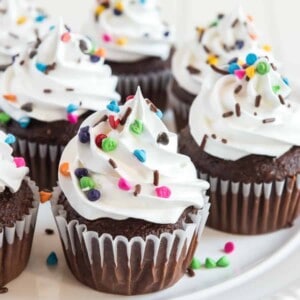 A plate of chocolate cupcakes with white swirled frosting without butter, topped with colorful sprinkles and chocolate shavings, displayed on a white surface.