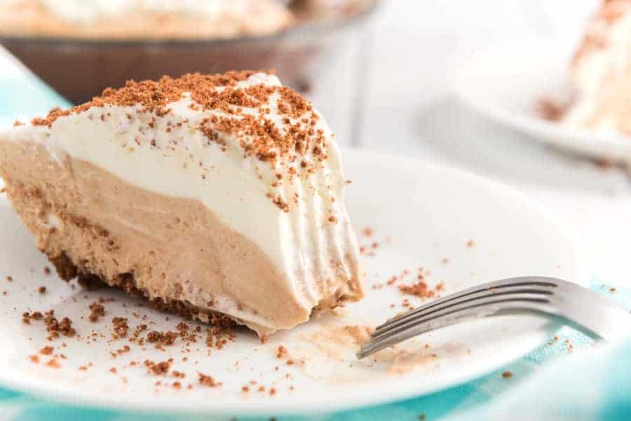 A slice of creamy chocolate peanut butter pie with layers of decadent chocolate and vanilla filling topped with a layer of whipped cream and sprinkled with crumbled chocolate on a white plate. A fork rests beside the slice, and another pie is blurred in the background.