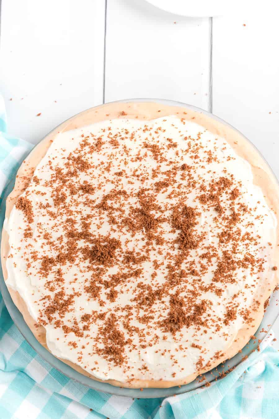A pie topped with fluffy icing and chocolate shavings sits on a table. The crust appears golden brown, adding to its allure. A light blue and white checkered cloth is partially visible to the side, enhancing the cozy ambiance.
