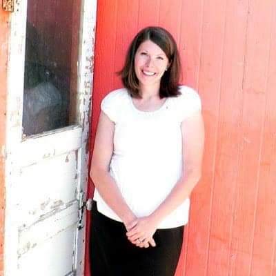 A brown-haired woman wearing a white shirt standing in front of a rustic red wall