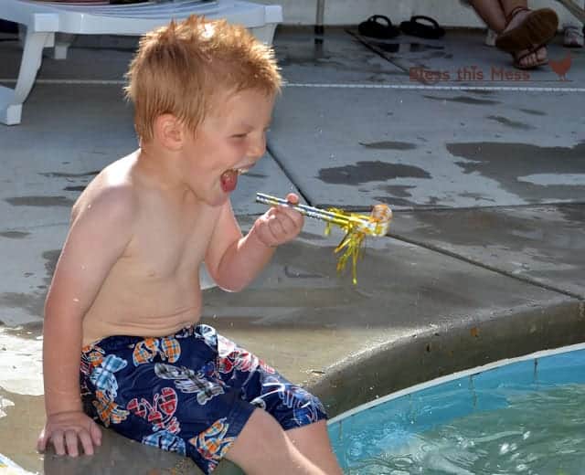 A little boy sitting with his feet in a pool, smiling and holding a party blower