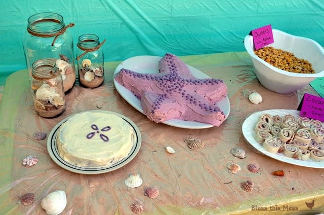 A party table with sand and shell decorations, a sand dollar cake, and a starfish cake