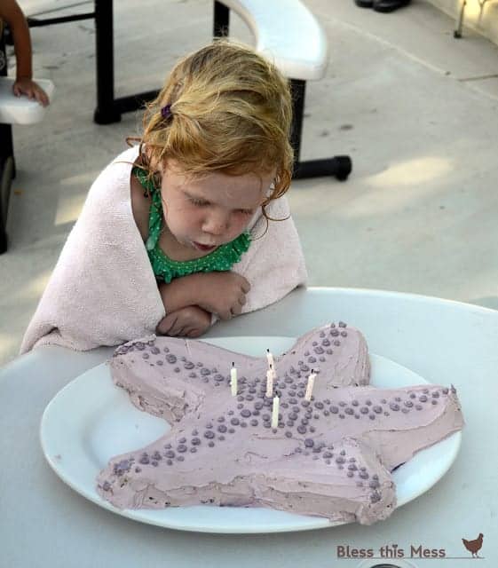 A girl in a bathing suit and towel blowing out candles on a starfish shaped birthday cake