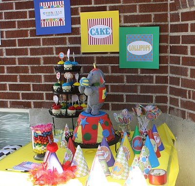 An elephant birthday cake on a table with circus-themed decorations, party hats, cupcakes in front of a brick wall with circus-themed signs