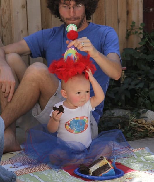 A baby wearing a blue tutu and circus-themed shirt and party hat sitting on the ground eating birthday cake