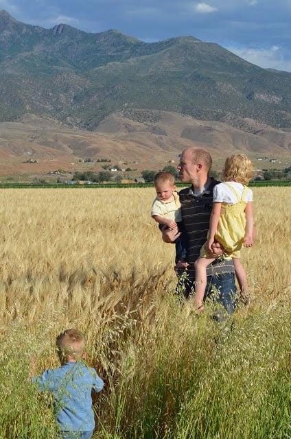 A man standing in long grass with mountains behind holding two young children and a third child is walking through the grass