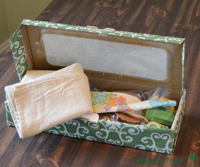 A box covered with green and white print filled with contents of a tea set, cloth napkins, and cookies