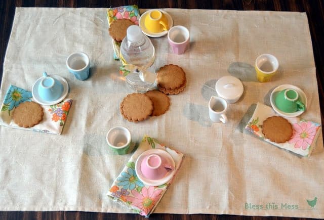 Four colorful tea cups and saucers with floral cloth napkins and a stack of cookies in the center