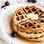 A stack of cherry waffles topped with two small butter slices and drizzled with syrup sits on a white plate. Fresh cherries and a white cloth with black print complement the scene on the marble surface.