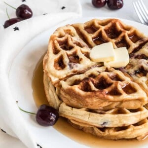 A stack of cherry waffles topped with two small butter slices and drizzled with syrup sits on a white plate. Fresh cherries and a white cloth with black print complement the scene on the marble surface.