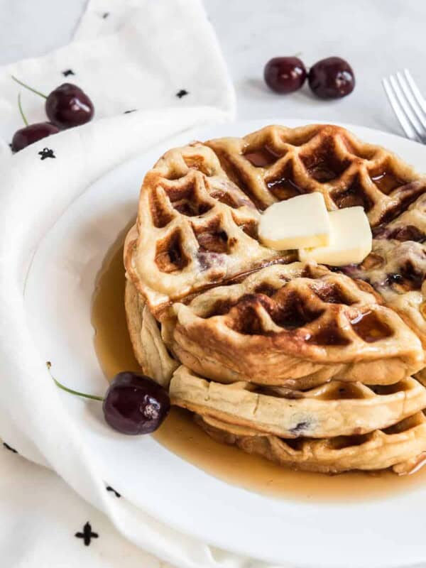 A stack of cherry waffles topped with two small butter slices and drizzled with syrup sits on a white plate. Fresh cherries and a white cloth with black print complement the scene on the marble surface.