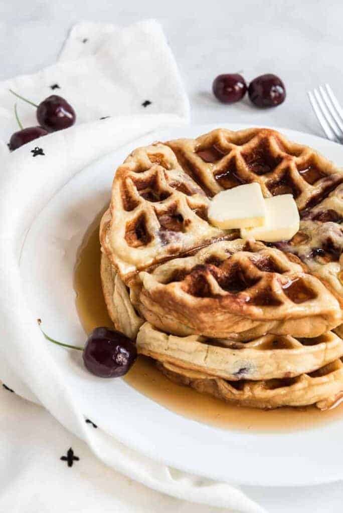 A stack of cherry waffles topped with two small butter slices and drizzled with syrup sits on a white plate. Fresh cherries and a white cloth with black print complement the scene on the marble surface.