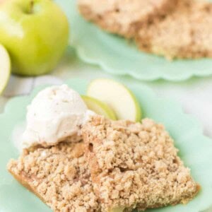 Two crumbly apple dessert bars, reminiscent of apple crumb cake, sit on a green plate with vanilla ice cream and apple slices. Another plate with more bars is in the background, alongside a whole green apple.