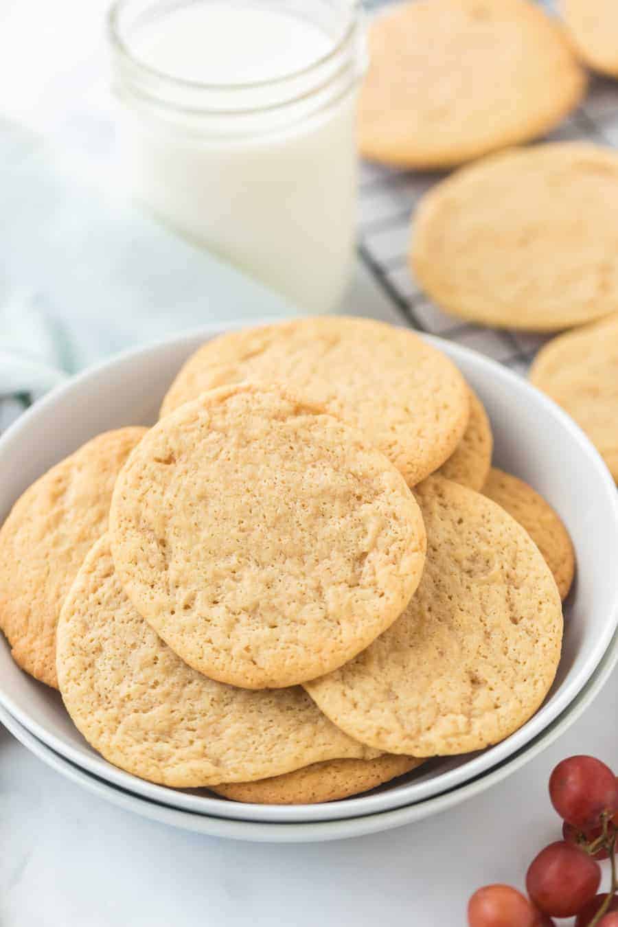 In the foreground is a bowl of cookies with some grapes in the bottom right-hand corner. In the background is a jar of milk and a cooling rack with cookies on it.