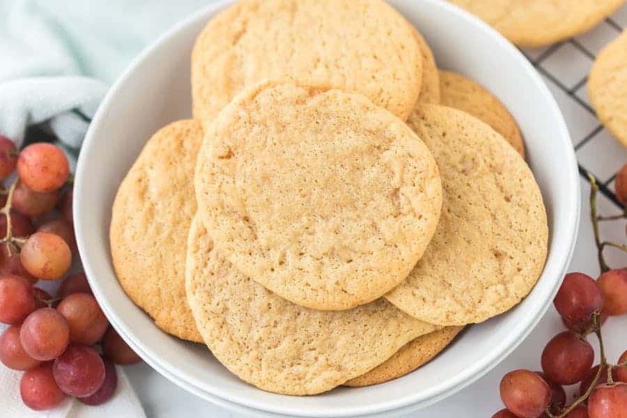 A bowl with baked cookies in it, surrounded by purple grapes and a cooling rack with more cookies on it.