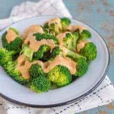 green broccoli florets on a white plate with cheese sauce on top sitting on black and white towel on a blue background.