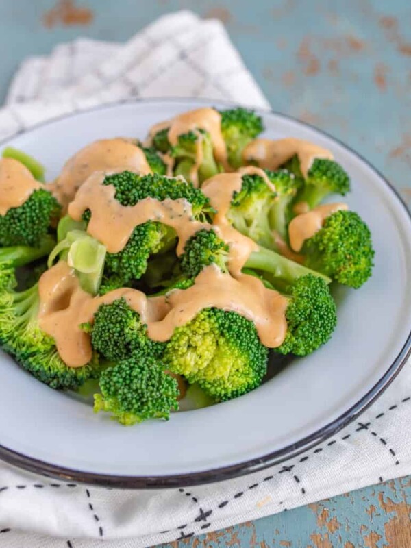 green broccoli florets on a white plate with cheese sauce on top sitting on black and white towel on a blue background.