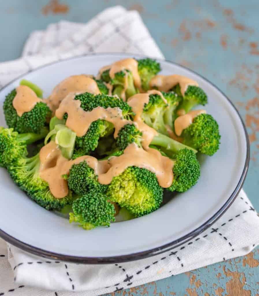 green broccoli florets on a white plate with cheese sauce on top sitting on black and white towel on a blue background.