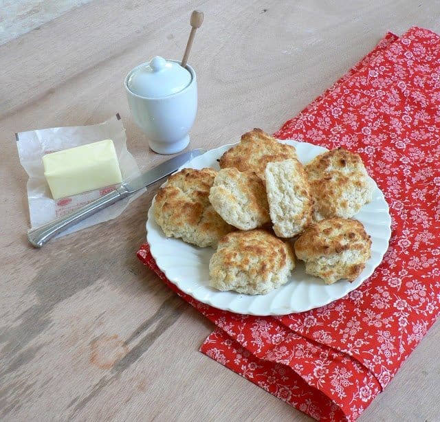 home made cream biscuits on white dish with red cloth under butter and knife and white dish beside it.