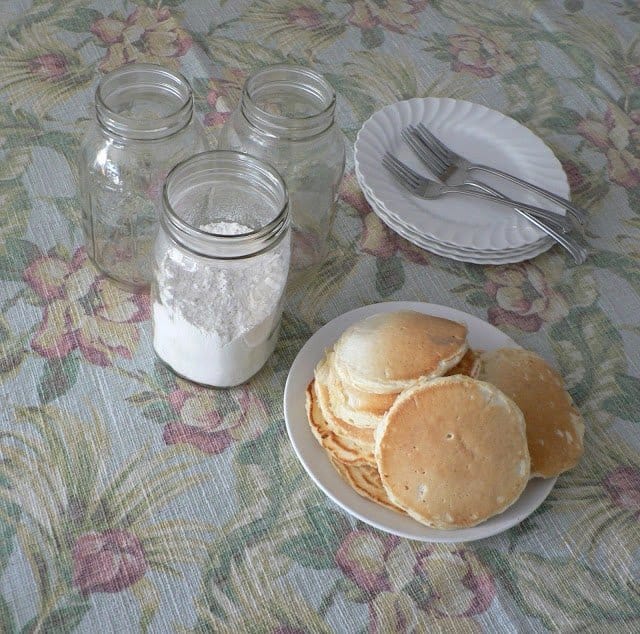 three mason jars with white plates, silver forks, and pancakes on a place on a flowered tablecloth.