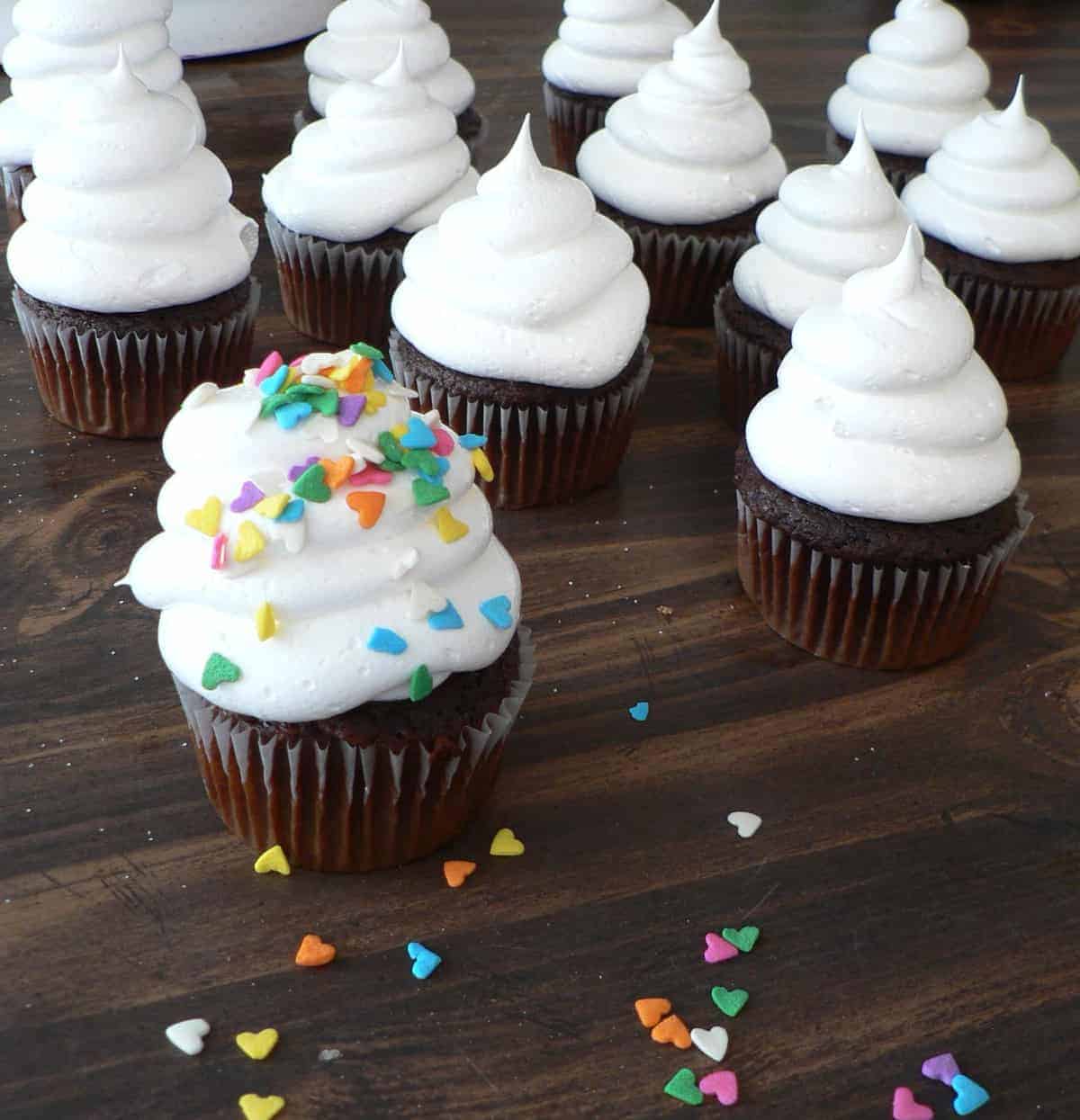 Chocolate cupcakes with tall swirls of white frosting without butter are arranged on a dark wooden surface. One cupcake in the front is topped with colorful heart-shaped sprinkles, and more sprinkles are scattered nearby.