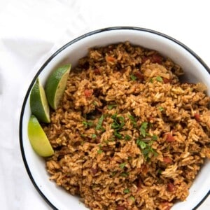A bowl of seasoned Spanish rice in rice cooker, garnished with fresh chopped herbs and served with lime wedges on the side, placed on a white surface with a folded white napkin nearby.