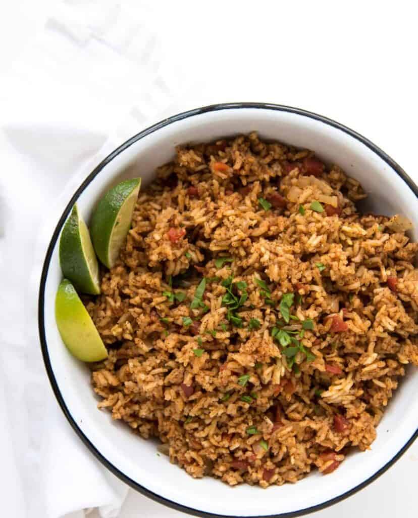 A bowl of seasoned Spanish rice in rice cooker, garnished with fresh chopped herbs and served with lime wedges on the side, placed on a white surface with a folded white napkin nearby.