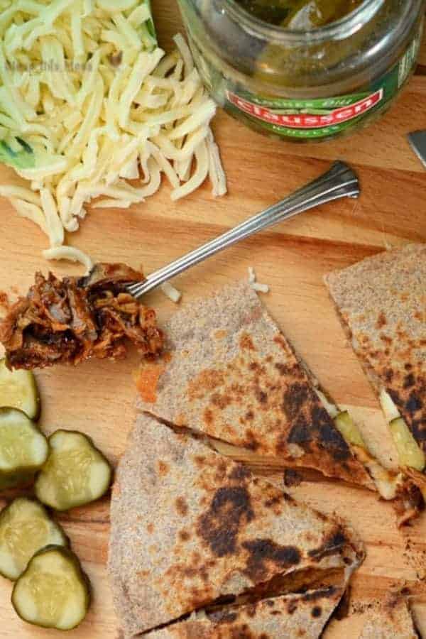 BBQ Pork Quesadillas on a cutting board with sliced pickles, shredded pork, and shredded cheese