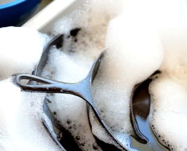 three black cast iron skillets in a sink full of soapy water.