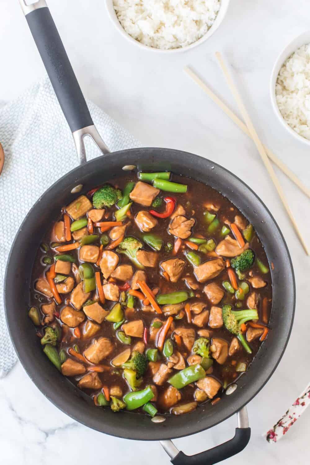 stir fry chicken and vegetables in a skillet next to chopsticks and bowls of white rice