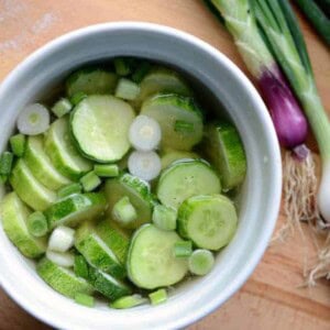 A white bowl brimming with quick pickled cucumbers and sliced green onions in a clear liquid. Fresh green onions, roots intact, lay beside the bowl on a wooden surface.