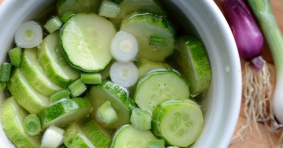 A white bowl brimming with quick pickled cucumbers and sliced green onions in a clear liquid. Fresh green onions, roots intact, lay beside the bowl on a wooden surface.