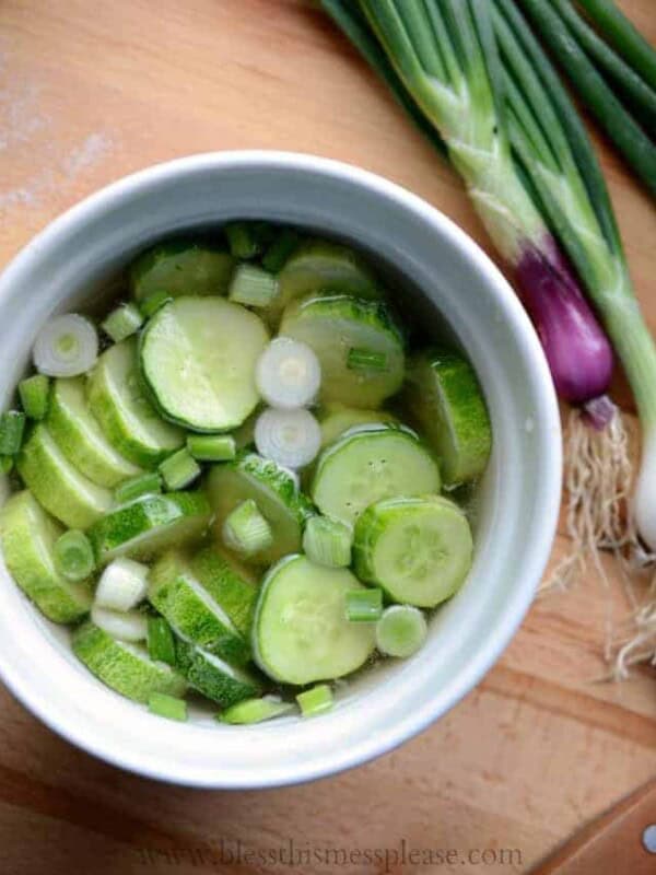 A white bowl brimming with quick pickled cucumbers and sliced green onions in a clear liquid. Fresh green onions, roots intact, lay beside the bowl on a wooden surface.